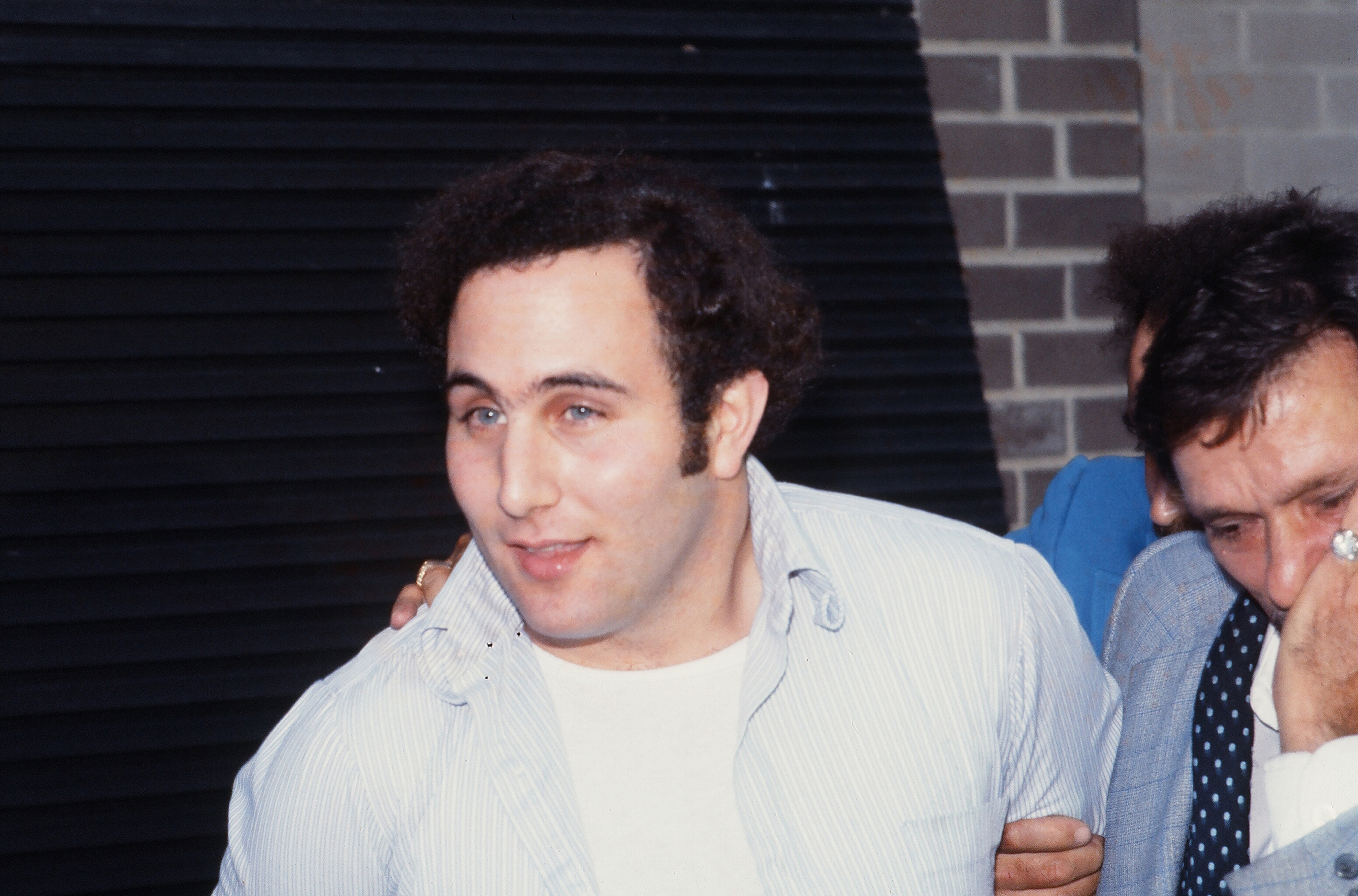 Police officers escort American accused (and ultimately convicted) serial killer David Berkowitz (left), known as the Son of Sam, into the 84th precinct station, New York, New York, August 10, 1977. (Image via Robert R. McElroy/Getty Images)